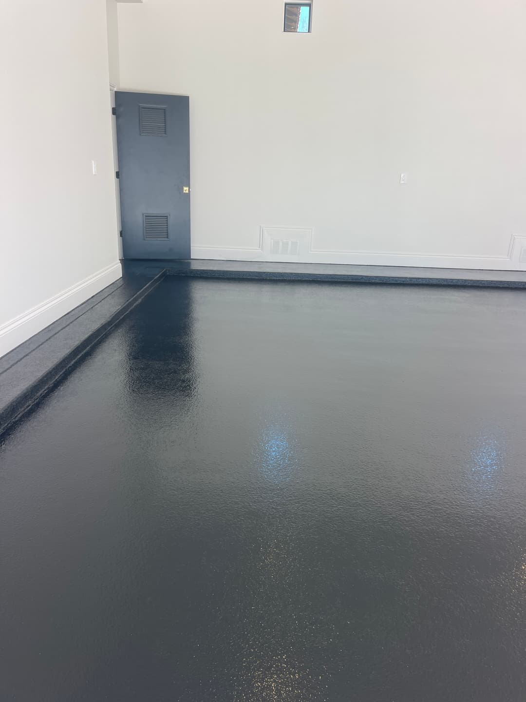 Glossy black epoxy floor in a modern, empty room with gray walls and a door.