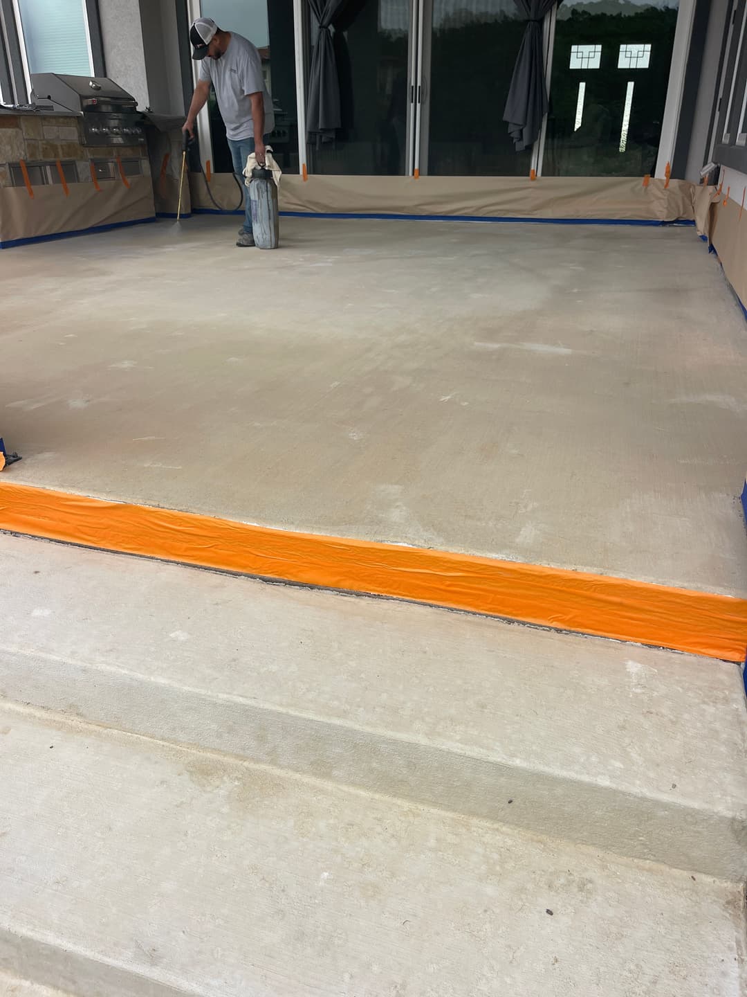 Worker applying sealant on a newly poured concrete patio with blue and orange safety barriers.
